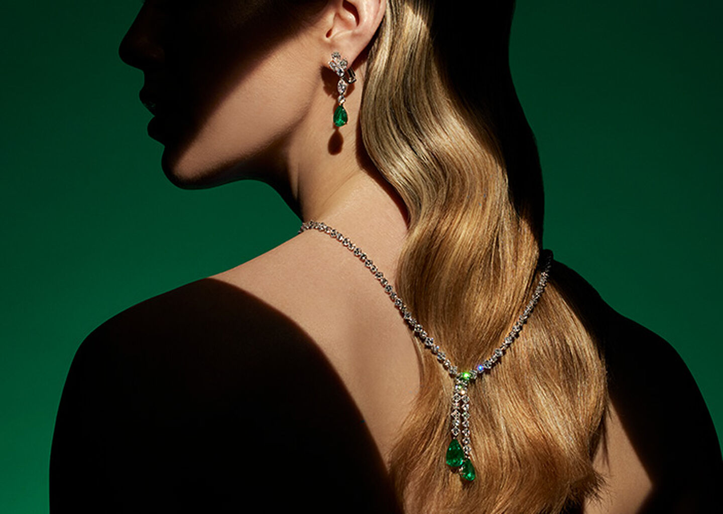 A woman wearing a Maison Birks emerald high jewellery necklace and earrings on a green background.